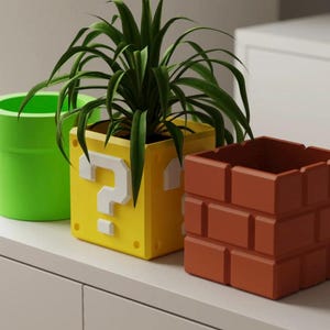 Könnte beinhalten: Drei farbenfrohe Pflanzgefäße in einer Reihe. Ein grüner Rohr-Pflanztopf, ein gelber Fragezeichen-Block-Pflanztopf und ein Pflanztopf im Ziegelstil. Der gelbe Pflanztopf hat ein weißes Fragezeichen auf der Vorderseite. Die Pflanzgefäße sind mit grünen Pflanzen gefüllt.