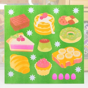 May include: A green square sticker sheet featuring colorful illustrations of various desserts. The sheet includes images of a brownie, pancakes, cookies, a pie slice, a donut, a custard, a croissant, a cupcake, and a piece of toast with bananas.