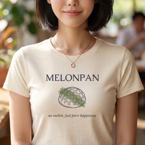 May include: A cream-colored short-sleeve t-shirt with a speckled texture. The front features the text MELONPAN in dark letters above a graphic of a melon pan bun with a green watercolor accent, followed by the text no melon, just pure happiness.