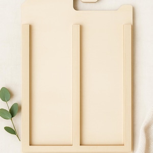 May include: A light beige wooden frame with a rectangular design, featuring a top hook and two vertical dividers. The frame is set against a neutral background with a sprig of green leaves and a white fabric.