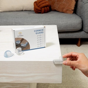 May include: A white box labelled "CORNER GUARD" with a blue and white graphic of corner protectors. Several clear corner protectors are on a light-coloured wooden table. A hand is holding a clear corner protector.