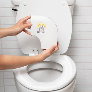 May include: A white plastic toilet seat attachment featuring a graphic of a bear inside a yellow sun and pink clouds with the text LUNIKO printed in black below the bear. The device is curved and designed to fit onto the rim of a standard toilet bowl.
