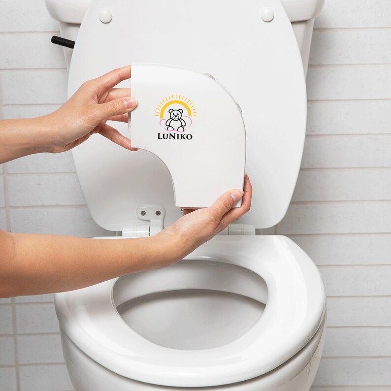 May include: A white plastic toilet seat attachment featuring a graphic of a bear inside a yellow sun and pink clouds with the text LUNIKO printed in black below the bear. The device is curved and designed to fit onto the rim of a standard toilet bowl.