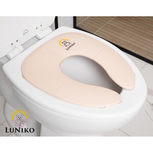 May include: A foldable, light beige toilet seat reducer placed on a white toilet. The seat features a small graphic of a teddy bear inside a yellow sun arch with the text LUNIKO printed below it. The design is smooth, rounded, and functional.