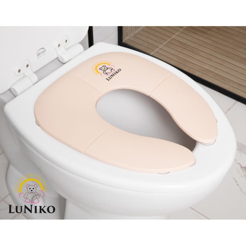 May include: A foldable, light beige toilet seat reducer placed on a white toilet. The seat features a small graphic of a teddy bear inside a yellow sun arch with the text LUNIKO printed below it. The design is smooth, rounded, and functional.