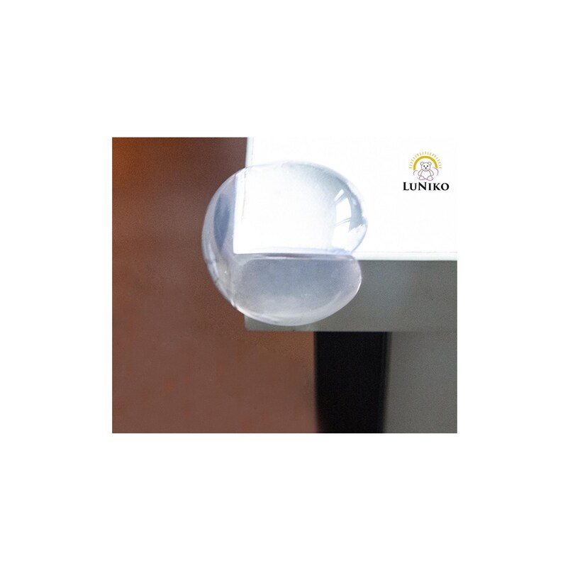 May include: A clear, rounded silicone corner protector attached to the edge of a white table. The protector is transparent and smooth, designed to cover sharp corners. The logo LUNIKO with a small bear icon is visible on the white surface.