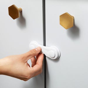 May include: A white plastic cabinet safety lock attached to two white cabinet doors. The cabinet doors feature two gold-colored hexagonal knobs. The locking mechanism is positioned horizontally across the gap between the two doors.