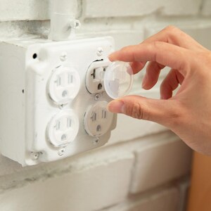 May include: A clear plastic socket plug cover being inserted into a white four-gang electrical wall receptacle. The receptacle is mounted on a white painted brick wall surface. The plug cover is circular with a small handle for easy removal.