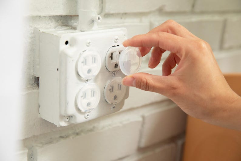 May include: A clear plastic socket plug cover being inserted into a white four-gang electrical wall receptacle. The receptacle is mounted on a white painted brick wall surface. The plug cover is circular with a small handle for easy removal.
