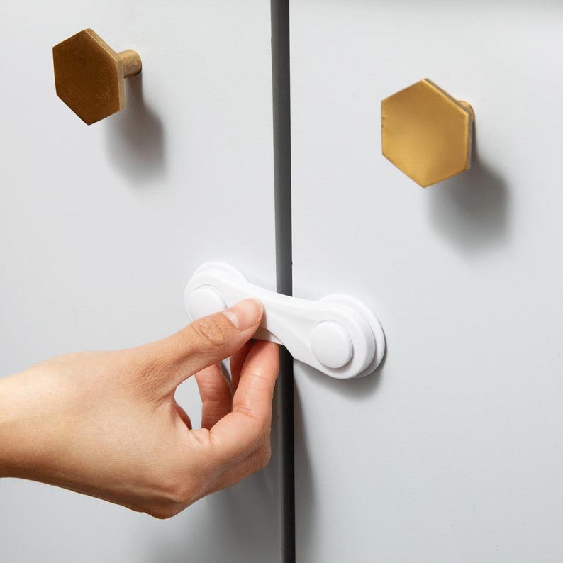 May include: A white plastic cabinet safety lock attached to two white cabinet doors. The cabinet doors feature two gold-coloured hexagonal knobs. The locking mechanism is positioned horizontally across the gap between the two doors.