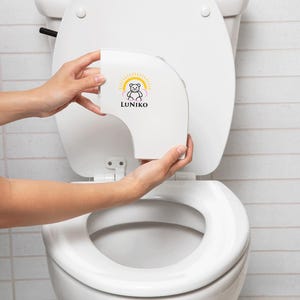 May include: A white plastic toilet seat attachment featuring a graphic of a bear inside a yellow sun and pink clouds with the text LUNIKO printed in black below the bear. The device is curved and designed to fit onto the rim of a standard toilet bowl.