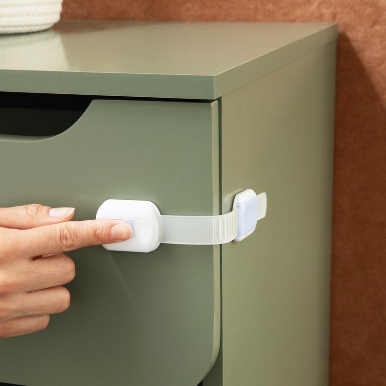 May include: A white plastic child safety lock with a flexible strap attached to the side of a sage green cabinet drawer. The locking mechanism features a central button and is secured to the drawer front and the adjacent cabinet frame.