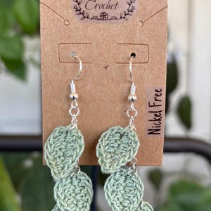 May include: Handmade green crochet leaf earrings with silver-colored hooks. The earrings are displayed on a brown tag with the text "True Crochet" and "Nickel Free". The leaves are a soft green color.