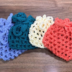 May include: Four handmade crochet bags in shades of blue, teal, cream, and coral. Each bag features a drawstring closure and a textured, open weave design. The bags are arranged on a wooden surface.