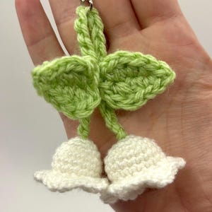 May include: A handmade crochet keychain featuring a lily of the valley design. The keychain has two white bell-shaped flowers and green leaves. The leaves and flowers are connected by a silver chain.