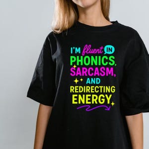 May include: Black t-shirt featuring the phrase "I'm fluent in phonics, sarcasm, and redirecting energy." The text is in green, purple, and yellow. The shirt has short sleeves and a round neckline.