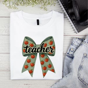 May include: White t-shirt featuring a large bow design with red apples on a green background. The word "teacher" is written across the bow in a black script font. The shirt is folded on a light wood surface.