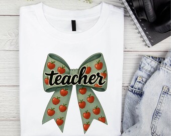 May include: White t-shirt featuring a large bow design with red apples on a green background. The word "teacher" is written across the bow in a black script font. The shirt is folded on a light wood surface.