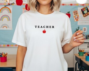 May include: White t-shirt featuring the word "TEACHER" in black, with a red apple graphic. The shirt has a crew neck and short sleeves. The background includes classroom decorations.