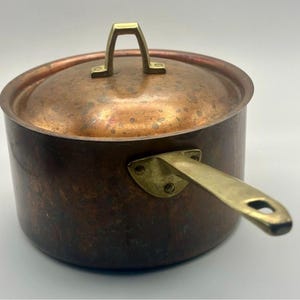 May include: A round copper saucepan with a matching lid, featuring a polished brass handle and a brass lid knob. The exterior of the pot and lid displays a weathered, dark copper patina with visible spots and natural color variations.