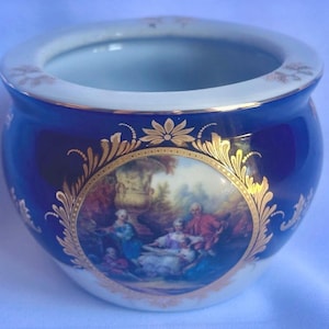 May include: A round porcelain bowl featuring a deep cobalt blue exterior with a central circular medallion depicting a group of figures in a garden setting. The bowl is accented with ornate gold filigree detailing and a white interior rim.