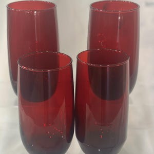 May include: Four red-tinted drinking glasses. Two are taller and more slender, while the other two are shorter and wider. The glasses have a smooth, rounded shape and a deep red color, perfect for serving beverages.