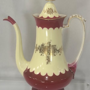 May include: A cream-colored porcelain coffee pot with a burgundy scalloped design. The pot features gold floral accents and a matching lid with a gold finial. The handle and spout are cream-colored with burgundy accents.