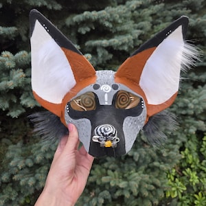 May include: A handcrafted animal mask featuring large pointed ears with white fur accents, a grey and black patterned face, and spiral designs on the eyes and nose. A silver safety pin with small beads is attached to the snout area.