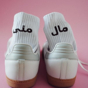 May include: A pair of white sneakers with light gray accents and brown soles. White socks with black Arabic text are visible inside the shoes. The background is a solid pink.