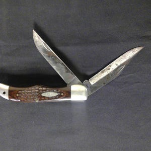 Puede incluir: Una navaja de bolsillo vintage con dos hojas abiertas. El mango es de color marrón oscuro con un patrón texturizado y detalles plateados. Las hojas son plateadas y muestran signos de desgaste.