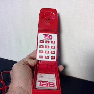 May include: A red Tab Flip Phone with a white keypad and red buttons. The phone has a flip-up design and includes the text "Tab" and "Flip Phone". The phone is held open, revealing a section for emergency contact information.