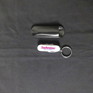 May include: A white multi-tool pocket knife keyring featuring the red Budweiser logo and the text King of Beers, accompanied by a black protective slip case. The multi-tool includes a metal keyring attached to one end of the white handle.