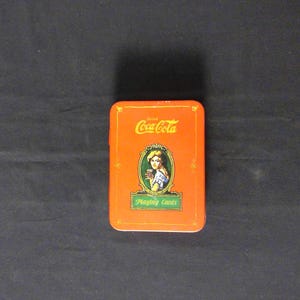 Puede incluir: Una caja rectangular naranja de cartas de juego Coca-Cola. La caja presenta el logotipo de Coca-Cola y una ilustración vintage de una mujer sosteniendo un vaso de Coca-Cola. Las palabras "Playing Cards" están debajo.