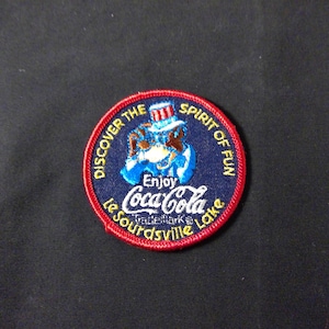 Peut inclure: Écusson brodé Coca-Cola avec un bord rouge et un centre bleu marine. Il présente un personnage de dessin animé portant un chapeau haut de forme et le texte "Enjoy Coca-Cola Le Sourdsville Lake". Les mots "Discover the Spirit of Fun" sont également inclus.