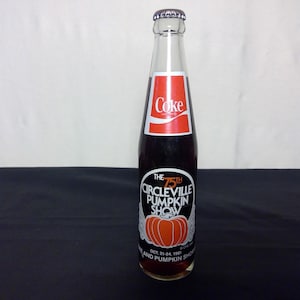 May include: A vintage Coca-Cola bottle with a red and white label. The bottle features a label for the 75th Circleville Pumpkin Show, with an orange pumpkin graphic. The bottle is clear, and the liquid inside is dark.