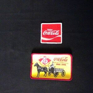 Może przedstawiać: Dwie naszywki Coca-Cola w stylu vintage. Jedna to czerwony kwadrat z logo Coca-Cola i napisem "Enjoy". Druga to prostokątna naszywka z wozem konnym, logo Coca-Cola i napisem "Delicious and Refreshing 1901-1976".