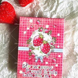 May include: A pink box with a strawberry design and the text "Strawberry Forest Souls Lenormand." The box features illustrations of strawberries, flowers, and a blue bow. Two large red strawberries are visible.