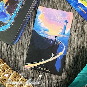 May include: A tarot card featuring a person standing on a cliff overlooking the ocean, with a lighthouse in the distance. The card has a colorful sky with the words "THE FOOL" at the bottom. A black box with a colorful design is also visible.