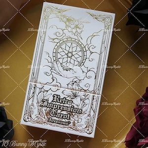 Könnte beinhalten: Eine weiße Tarotkartenbox mit goldenen floralen und geometrischen Designs. Auf der Box steht "Retro Impression Tarot" mit "Arcanums" darunter. Die Oberseite der Box zeigt eine Hand, die ein sternförmiges Rad hält.
