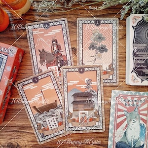 K&ouml;nnte beinhalten: Ein Set Lenormand-Orakelkarten in gedeckten Pfirsich- und Braunt&ouml;nen mit kunstvollen R&auml;ndern. Die Karten zeigen Illustrationen eines Samurai zu Pferd, eines Kleeblatts, eines Schiffes auf Wellen und eines Hauses sowie ein Begleitheft mit einem Fuchs.