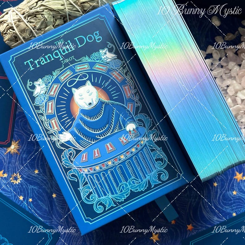 May include: A blue tarot deck box titled The Tranquil Dog Tarot featuring a central illustration of a dog in a robe at a table, accompanied by a deck of cards with iridescent holographic edges and a blue and gold star patterned surface.