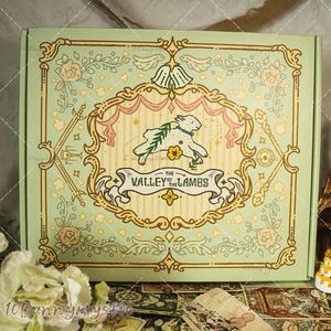 May include: A decorative box with a light green background and a central design featuring a lamb, flowers, and the text "The Valley of the Lambs." The box has a vintage aesthetic with ornate floral and scrollwork borders.