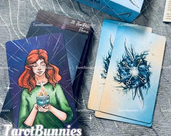May include: A deck of Tarot cards with a box labeled "Nest of Light." The cards feature illustrations of a woman with red hair and a candle, and abstract blue and white designs. The brand name "TarotBunnies" is visible.