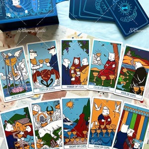 May include: A tarot card deck featuring white dog illustrations on each card. The set includes a blue storage box and ten cards labelled Ace through Ten of Cups, displaying various scenes with dogs, cups, and landscapes in a colourful graphic style.