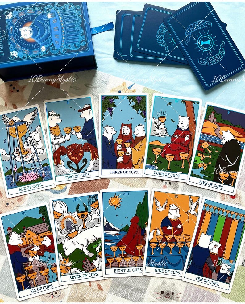 May include: A tarot card deck featuring white dog illustrations on each card. The set includes a blue storage box and ten cards labelled Ace through Ten of Cups, displaying various scenes with dogs, cups, and landscapes in a colourful graphic style.