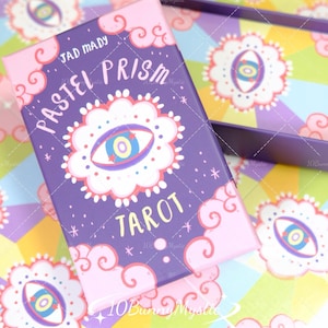 Op de afbeelding: Een paarse tarotkaartdoos met de woorden "Pastel Prism Tarot" en een oogontwerp. De doos heeft roze accenten en is omgeven door een kleurrijke achtergrond met vergelijkbare oogontwerpen. De merknaam "Jad Mady" is ook zichtbaar.