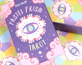 May include: A purple tarot card box with the words "Pastel Prism Tarot" and an eye design. The box has pink accents and is surrounded by a colourful background with similar eye designs. The brand name "Jad Mady" is also visible.