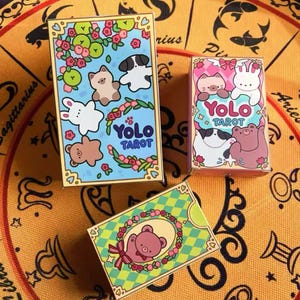 Könnte beinhalten: Drei farbenfrohe Tarotkartenboxen mit niedlichen Tierillustrationen. Die Boxen haben ein blaues, rosa und grünes Farbschema mit dem Text "Yolo Tarot". Die Illustrationen zeigen einen Bären, ein Kaninchen, eine Katze und einen Hund. Die Boxen befinden sich auf einer orangefarbenen Oberfläche mit Muster.