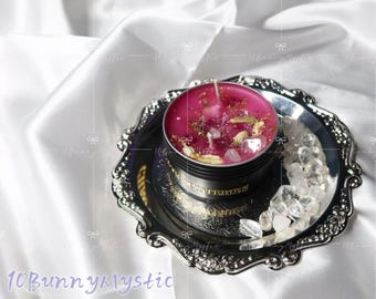 Puede incluir: Una vela rosa con hierbas secas y cristales en una lata negra sobre una bandeja decorativa plateada. La bandeja está adornada con cristales de cuarzo transparente y descansa sobre satén blanco. La lata de la vela tiene letras doradas.