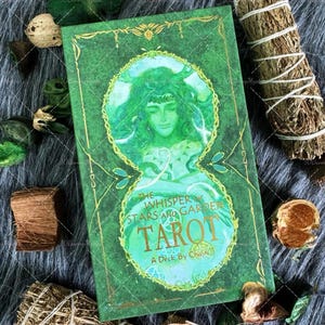 May include: A green tarot card deck titled "The Whisper of the Stars and Garden Tarot" with a watercolor illustration of a woman. The deck is surrounded by dried herbs and decorative elements on a gray fur-like surface.